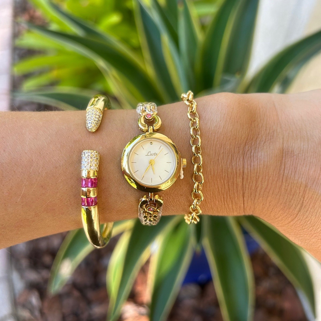 Gold watch and bracelets on a wrist with a plant in the background