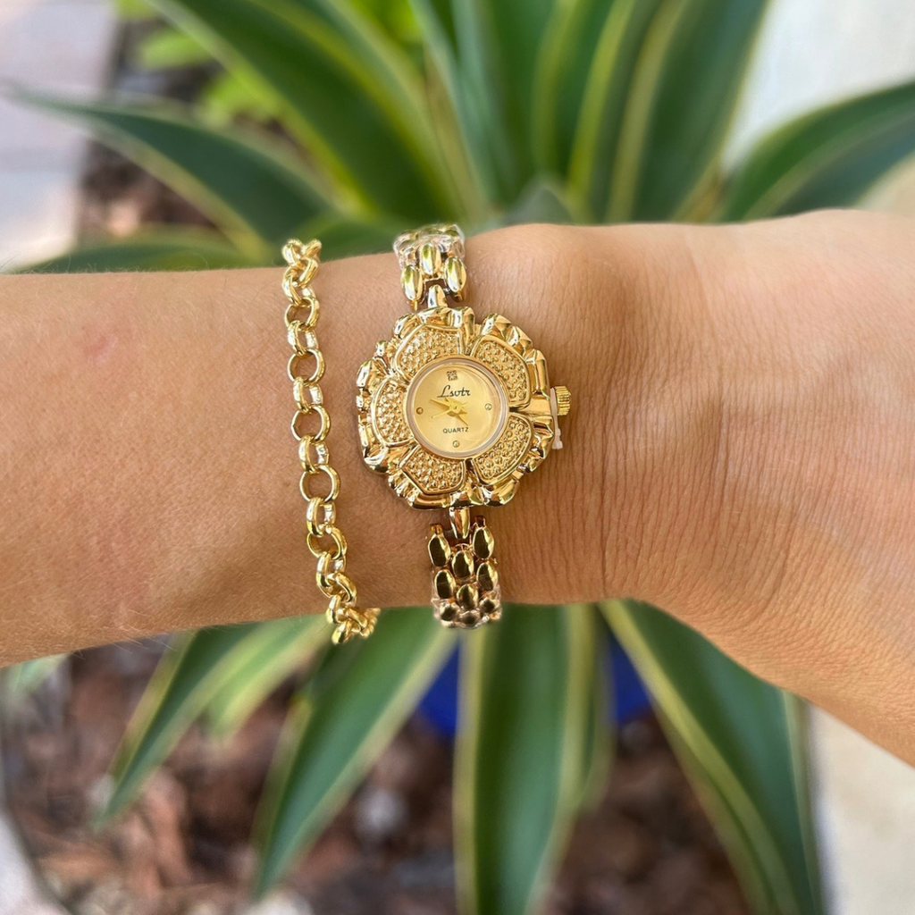 Gold watch and bracelet on a wrist with a plant in the background
