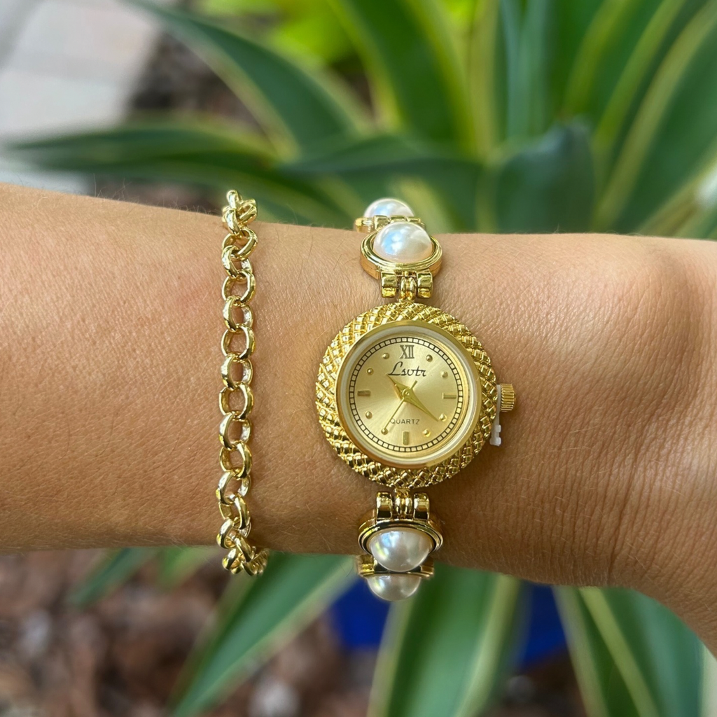 Gold wristwatch with pearl accents on a wrist, blurred green plants in the background