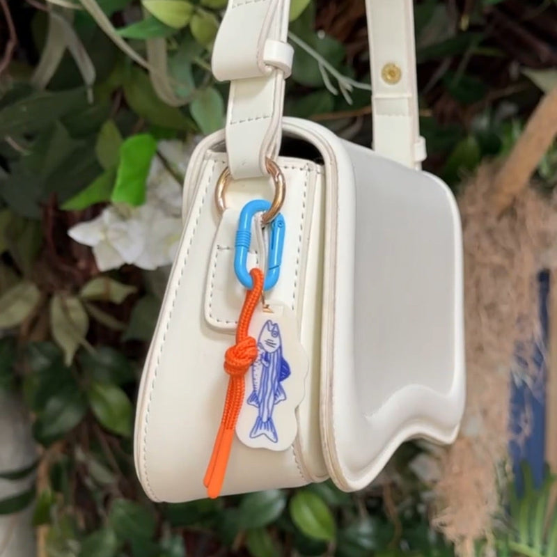 Blue acrylic fish carabiner bag charm with orange cord clipped onto white handbag
