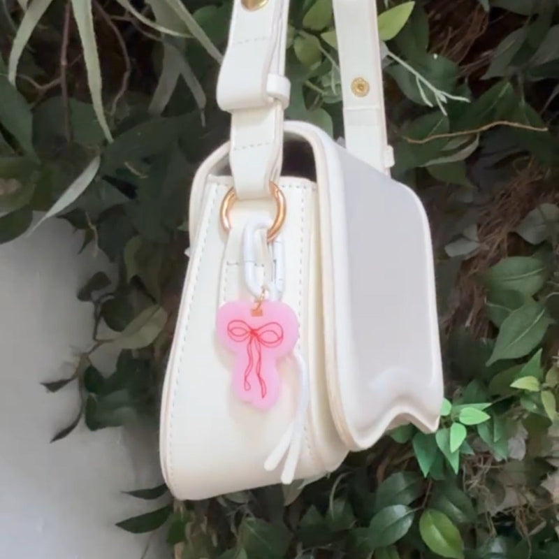 Pink acrylic bow carabiner bag charm with red bow detail clipped onto white handbag