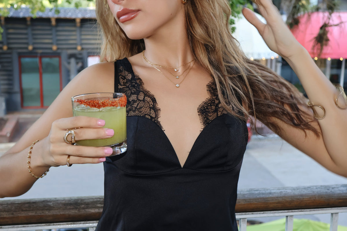 Woman in a black lace top wearing a gold bezel crystal solitaire necklace layered with gold chains, cuffs and rings